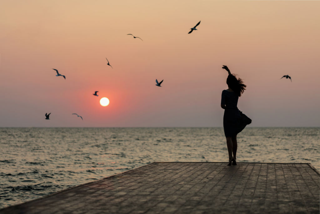Growing Our Spiritual Journey. Image of the silhouette of a woman with her hand raised, standing on a pier on the ocean. The sun is rising on the horizon and the silhouette of multiple birds can also be seen against the colorful sky.