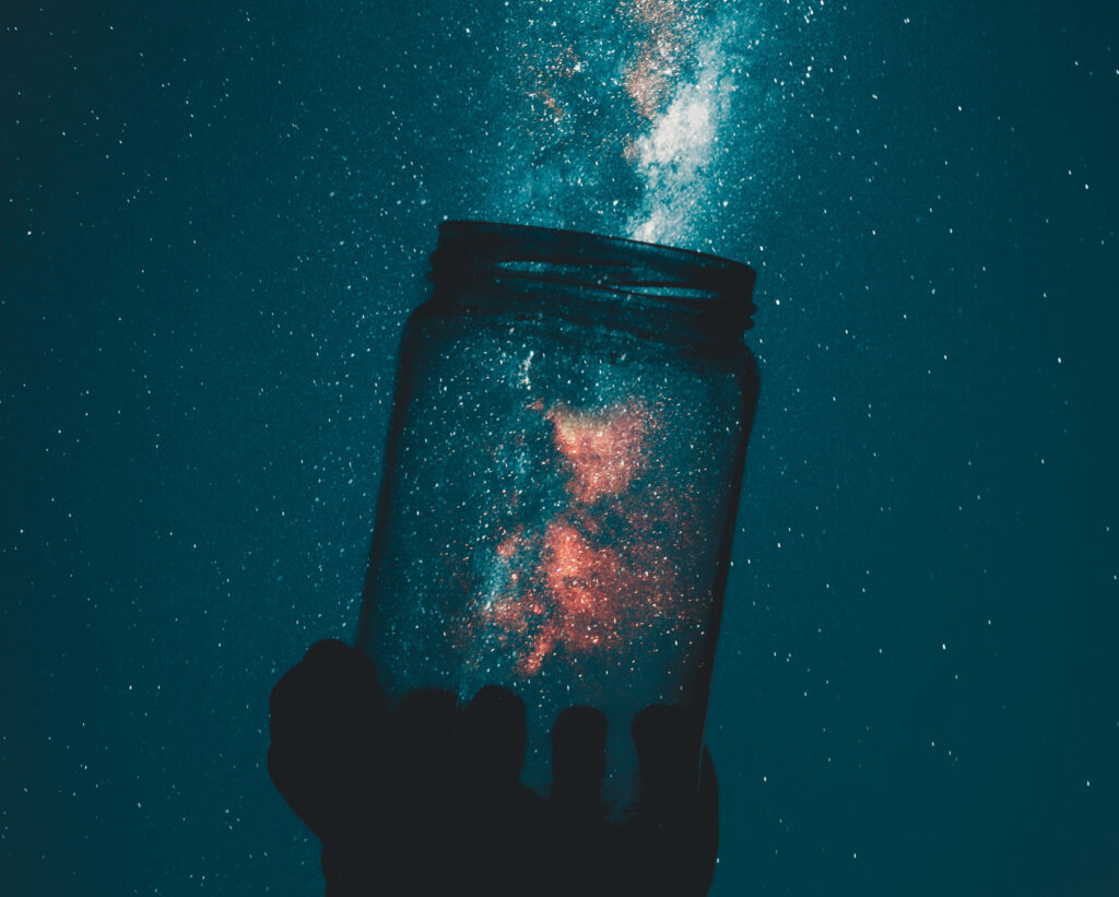 Receiving the Extraordinary. Image a person holing up a glass jar. A galaxy sky can be seen in the background through the glass jar. Joy.