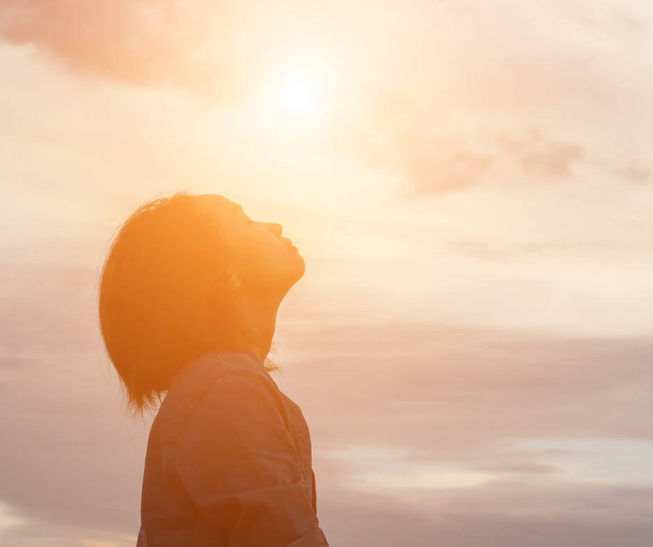 Self-Forgiveness as a Spiritual Practice. Image of a woman holding her head back and looking up toward the sky as sunlight shines over her face.