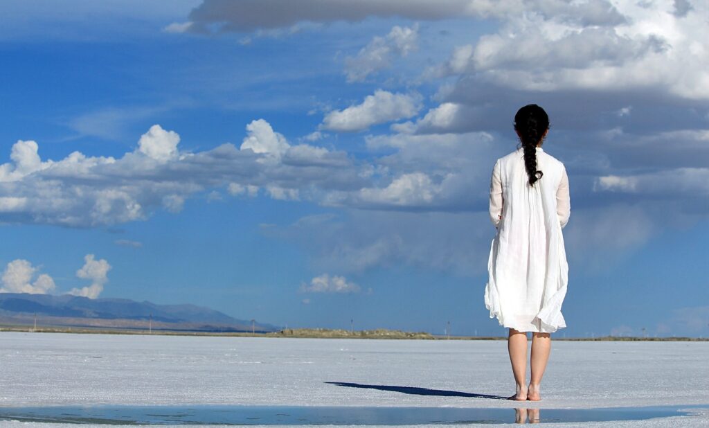 The Power of Looking Back. Self-reflection. Image of a woman in a white dress standing with her back facing the camera in an open, sandy area next to a puddle of water. In the background is a horizon line of mountains an a blue sky with some clouds.