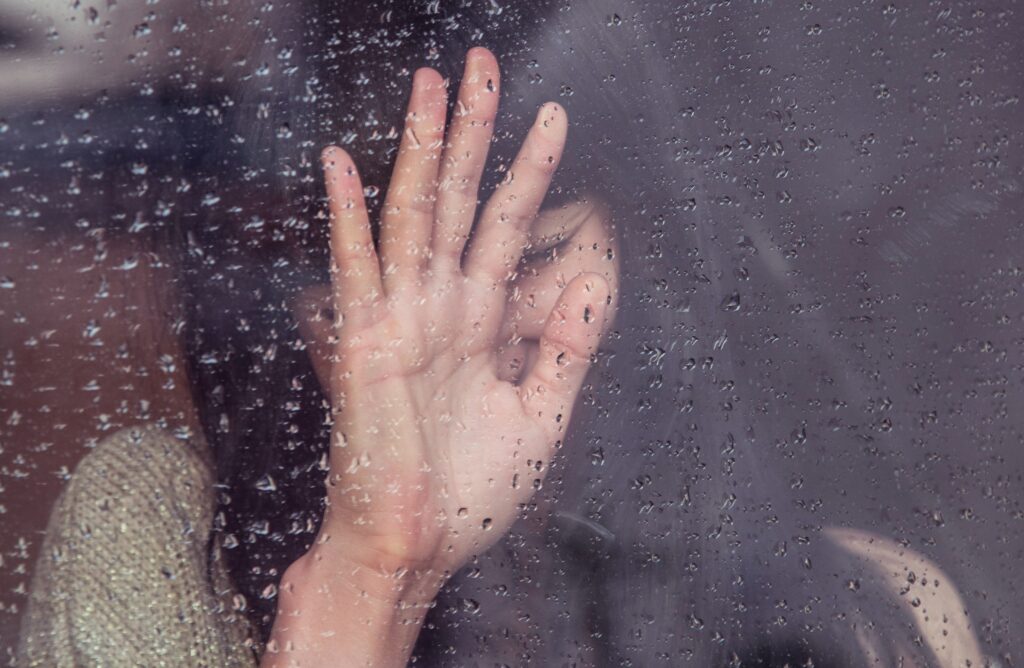 A Good Bad Day. Image of a woman covering her face by holding her hand up against a glass window covered in rain drops in front of her.