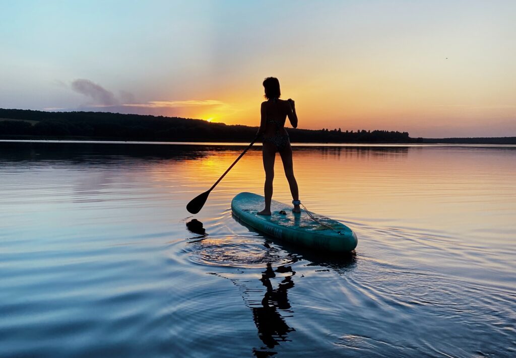 Freedom. Image of the silhouette of a woman holding a paddle, standing on a paddleboard on a lake. The sun and a forest of trees can be seen on the horizon in the background.